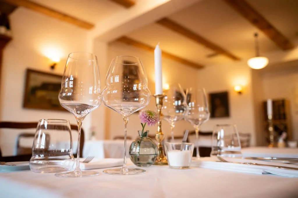 Im Landhaus Elise in Schildow bei Berlin sieht man einen geschmackvoll eingedeckten Tisch mit schön geschwungenen Weingläsern, einer dezenten Vase in der eine kleine Blume steht und im Hintergrund sieht man in warmem, goldenen Licht verschwommen den Gastraum. © Vanessa Bohneberg