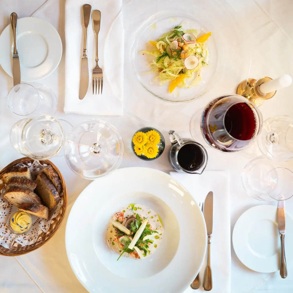 Auf einem eingedeckten Tisch im Landhaus Elise in Schildow bei Berlin sieht man aus der Vogelperspektive auf den Tisch auf dem Gläser mit Rot- und Weißwein stehen, ein Brotkorb mit frisch gebackenem Brot und Butter, zwei Tellern mit Vorspeisensalaten, Besteck, eine Vase mit gelben Blumen und eine weiße Kerze in einem goldenen Kerzenständer. © Vanessa Bohneberg