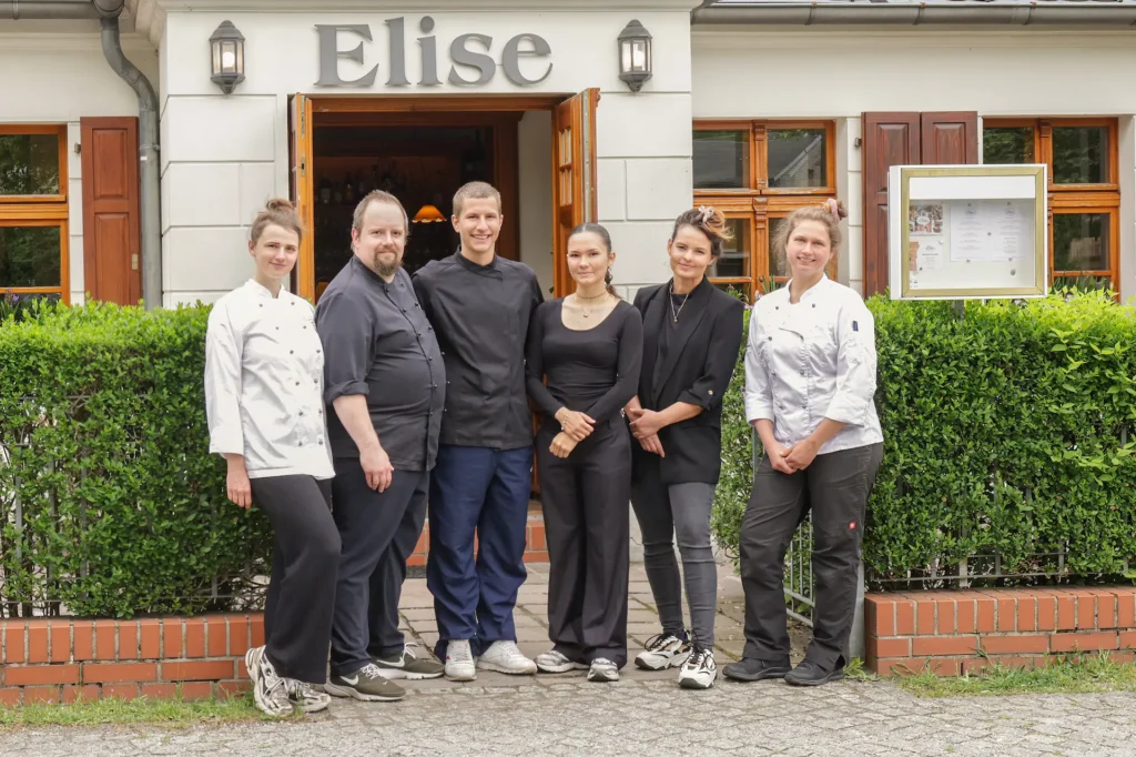 Auf dem Foto sieht man die Konditormeisterin Levke Adams und den Koch Sidney Adams mit ihrem Team des Landhaus Elise in Schildow bei Berlin vor dem Eingang zum Landhaus stehen. Man sieht sechs Personen nebeneinander, davon alle in ihrer Arbeitskleidung: Levke Adams, Sidney Adams, ihren Küchenhelfer, zwei Servicekräfte und eine weitere Konditormeisterin.